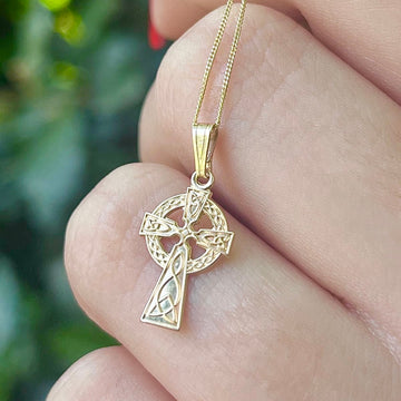 10k Gold Celtic Cross Necklace held on one hand and bushes in background.